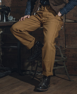 Brown double-knee canvas pants in a bent-leg stance, showing reinforced knee and side pocket.