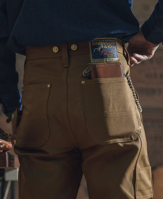 Brown canvas pants front with snap buttons, chestnut-toned tool loops, and utility pockets.